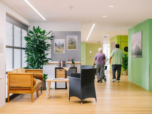 Zwei Personen gehen durch einen hellen Flur in einem Pflegeheim; vorne eine gemütliche Sitzecke mit Pflanzen und Bildern.